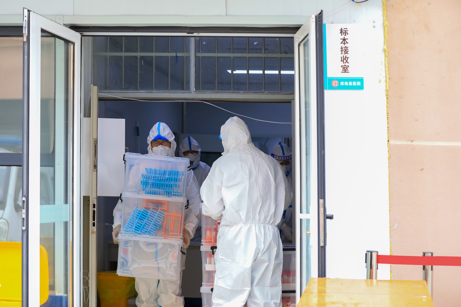 Two workers in white PPE carry boxes of supplies into a facility
